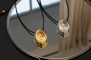 Two wax-seal pendants, gold and silver lion-head on black cords, resting on a round mirror with a silver rim.