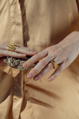 Close-up of hands wearing bold gold-coloured sculptural rings, left hand holding rough barnacled wood against beige fabric.