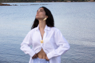 Model wearing a gold brooch pinned to a white buttoned shirt, blurred at waist-up against a calm waterside background.