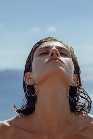 Model wearing large twisted gold hoop earrings in a low-angle close-up, sunlit by the sea; wet short dark hair.