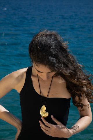 Young woman by the sea, wearing a large gold-coloured shell-shaped pendant on a black cord, hand gently touching the piece