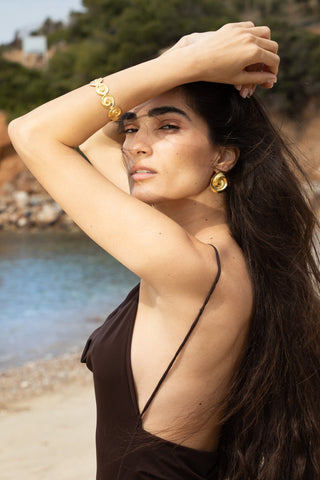 Woman by a rocky shore in a dark brown sleeveless top, arm raised, wearing large gold spiral earrings and matching bracelet.