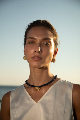 Close-up portrait of a woman wearing gold starburst earrings and a matching pendant on a dark beaded necklace, sunlit seaside.