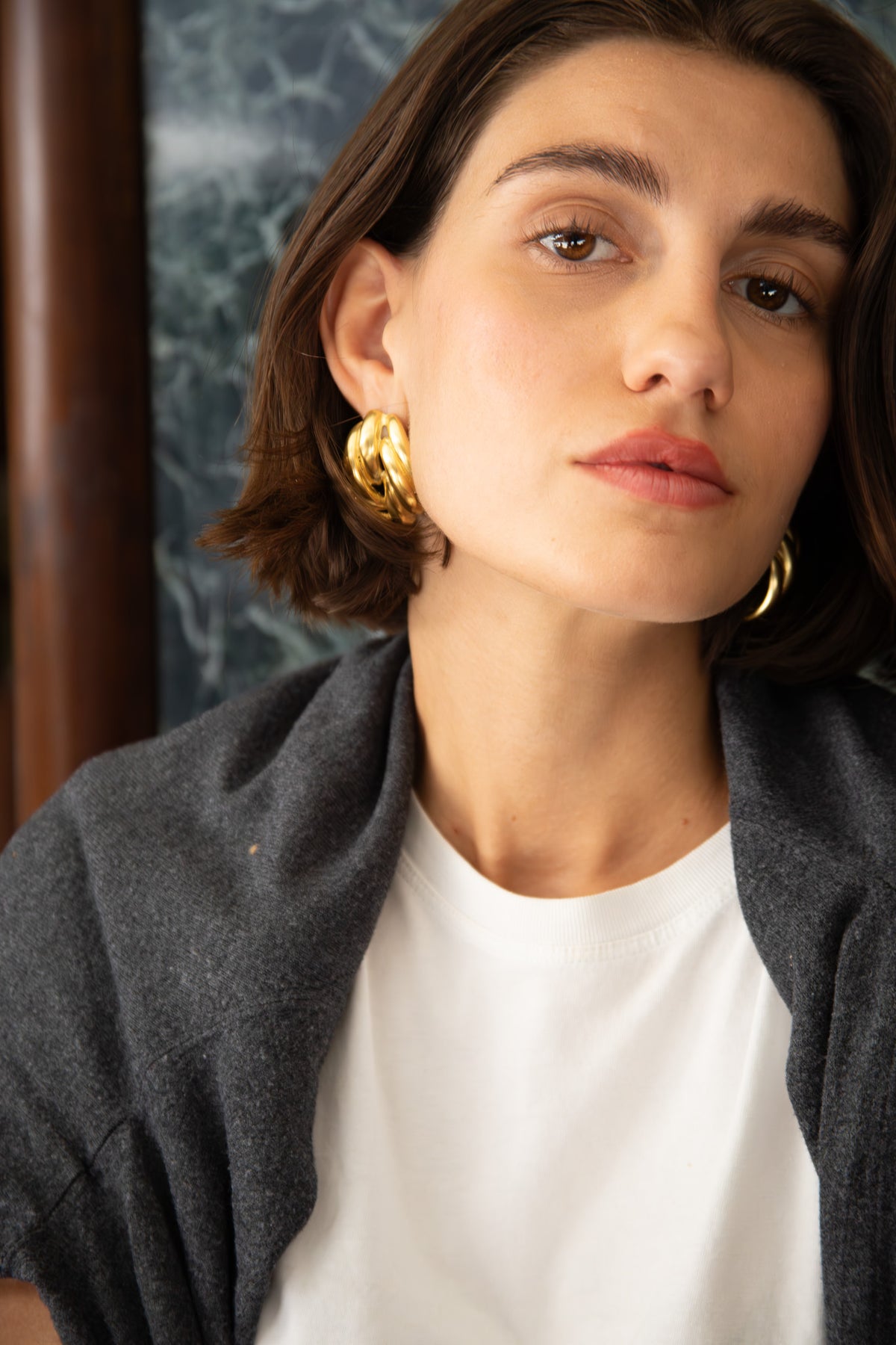 Close-up of young woman wearing large chunky gold hoop earrings, white crew-neck and draped dark grey sweater.