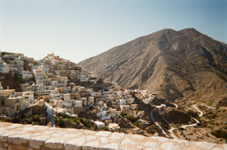 Densely built hillside village of white boxy houses rising towards a rocky mountain, stone wall in the foreground