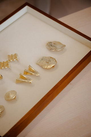 Five pairs of gold-coloured earrings (leaf, zigzag, cone, two intricate circular and square motifs) on a white tray — close-up.