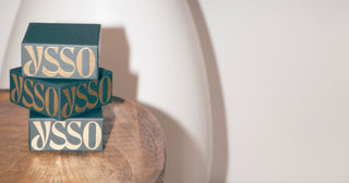 Stack of three dark teal YSSO jewellery boxes with gold lettering on a round wooden table, eye‑level and off‑centre
